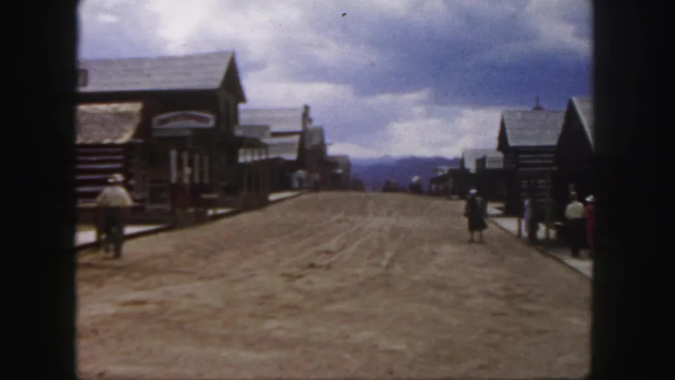 1958 Colorado 8mm view down a dusty Western-style street, with visitors spaced along boardwalks and log-front buildings on either side.