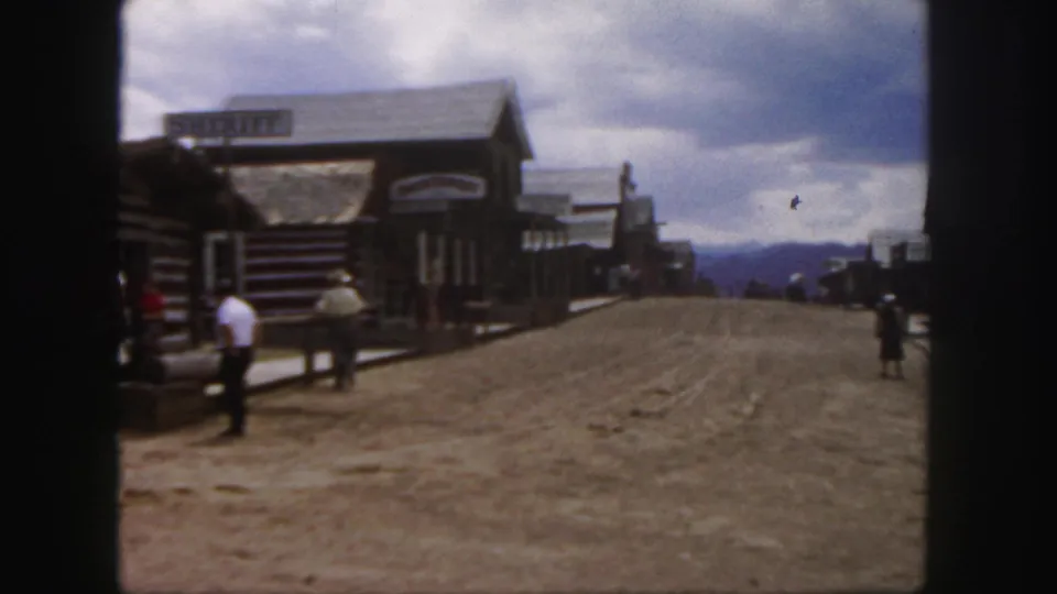1958 Colorado home-movie frame with a closer visitor in a white shirt, rustic storefronts, boardwalk edges, and a dusty Western-style street.