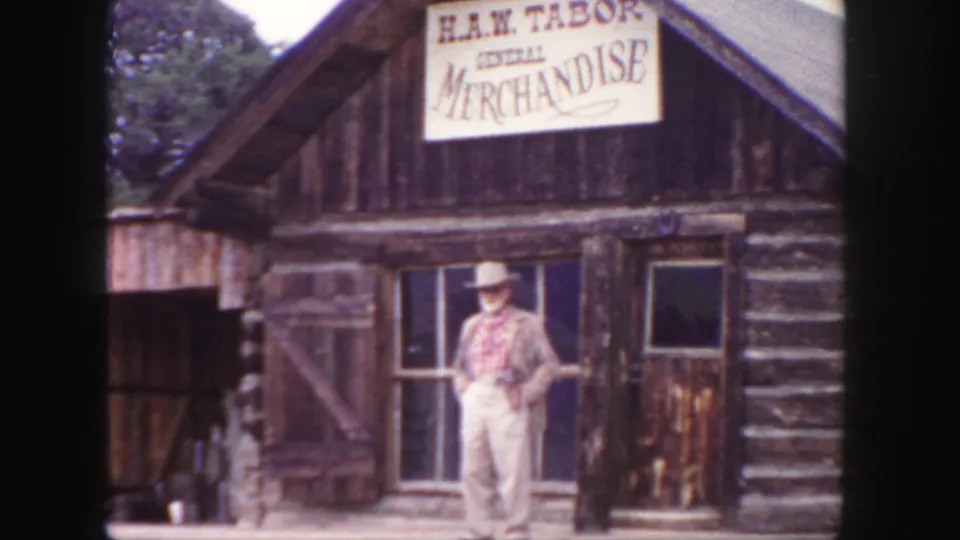 Man in cowboy hat and plaid shirt posed at the H.A.W. Tabor General Merchandise log building in Colorado, 1958.