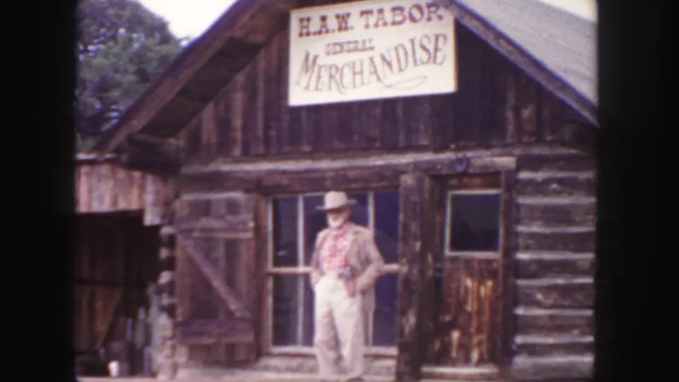 A western-dressed man stands at the log general store facade, with the H.A.W. Tabor sign visible above.