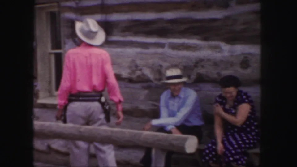 A cowboy-dressed man in a pink shirt walks away near two seated visitors in the same Western-style Colorado setting.