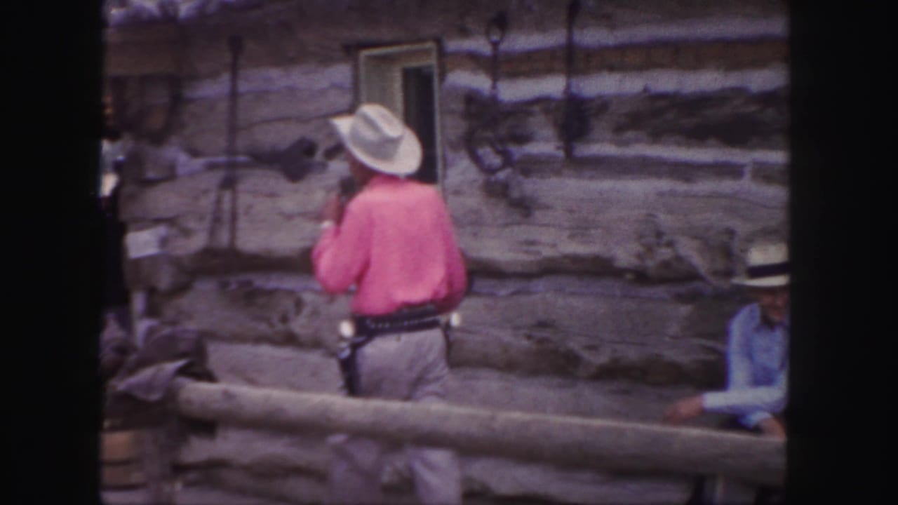 Cowboy in pink shirt and white hat stands by a wooden railing outside log buildings.