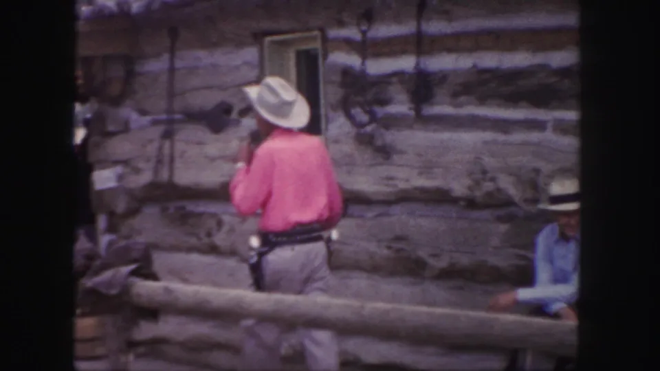 1958 Colorado home-movie frame showing a cowboy-costumed man in a pink shirt, white hat, and holsters near rustic log structures.