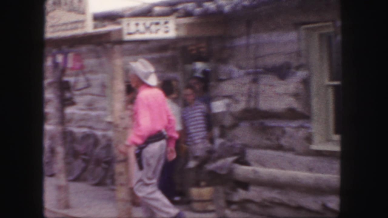 The cowboy-costumed man walks beside a rustic storefront with a partially visible sign and several visitors in the background.