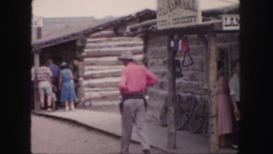 1958 Colorado frame with the pink-shirted cowboy walking past a rustic shop sign, wagon wheel, log building, and visitors on a dirt path.