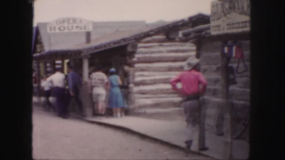 The cowboy-costumed man walks away through the Colorado Western-themed street as visitors gather near a log building and OPERA HOUSE sign.