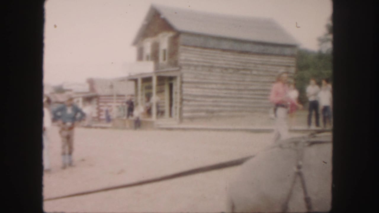 Cowboy-dressed man and other people near log buildings on a dusty street.