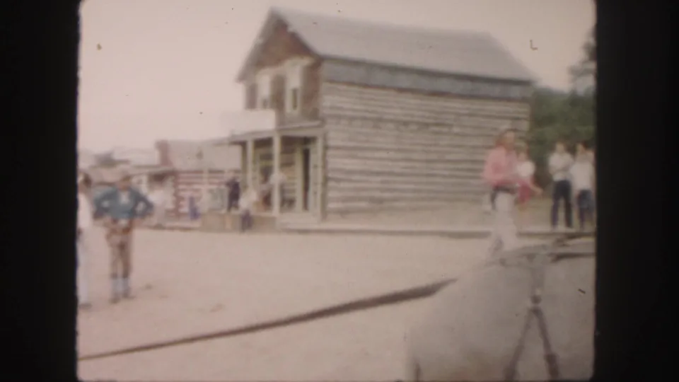1958 Colorado home-movie frame showing a cowboy-dressed man, pedestrians, and rustic log buildings along a dusty street.