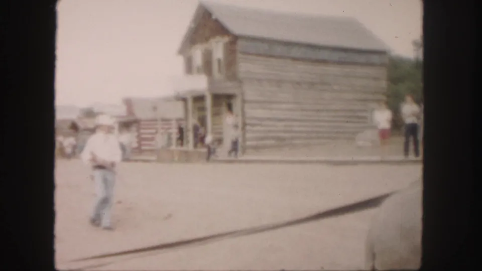 A man in a white hat crosses a dusty Colorado street with rustic log buildings and scattered visitors behind him.