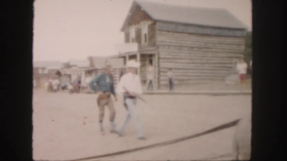 Two men, one in cowboy-style clothing and one in a white hat, walk near a log building in the 1958 Colorado street scene.