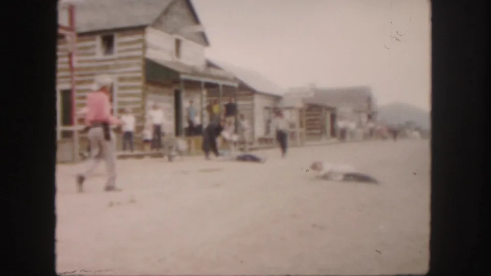 A cowboy-dressed man walks past a row of log storefronts as several people stand or move along the dusty street.
