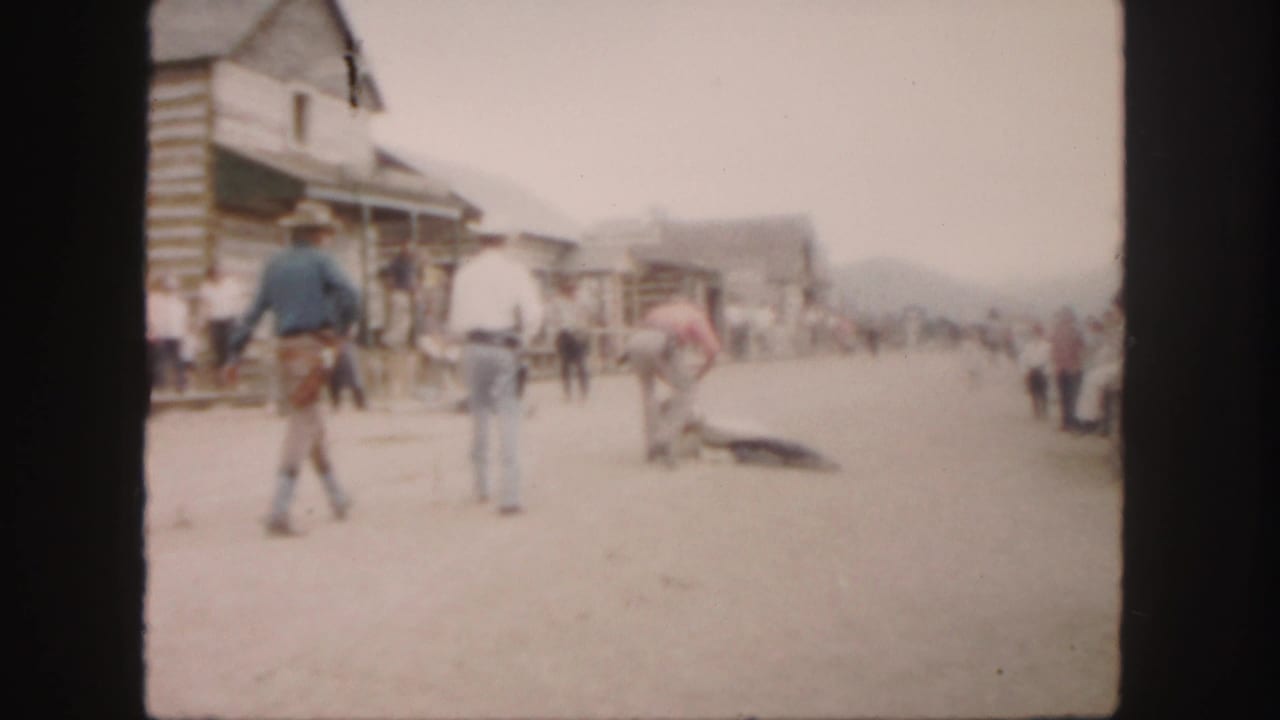 Men in cowboy hats stand on a dusty street near log-style buildings and an animal on the ground.
