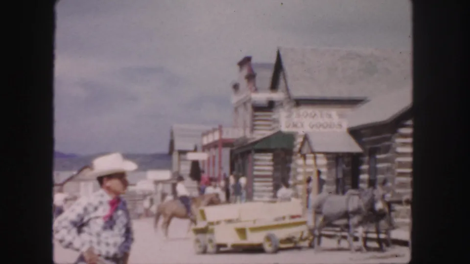 1958 Colorado home-movie frame with a man in a cowboy hat walking past Western storefronts, horses, a wagon, and visitors.