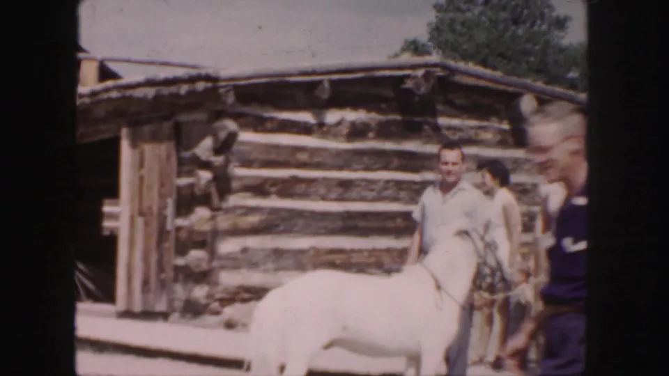 1958 Colorado frame showing visitors gathered near a white horse outside a rustic log cabin in bright daylight.
