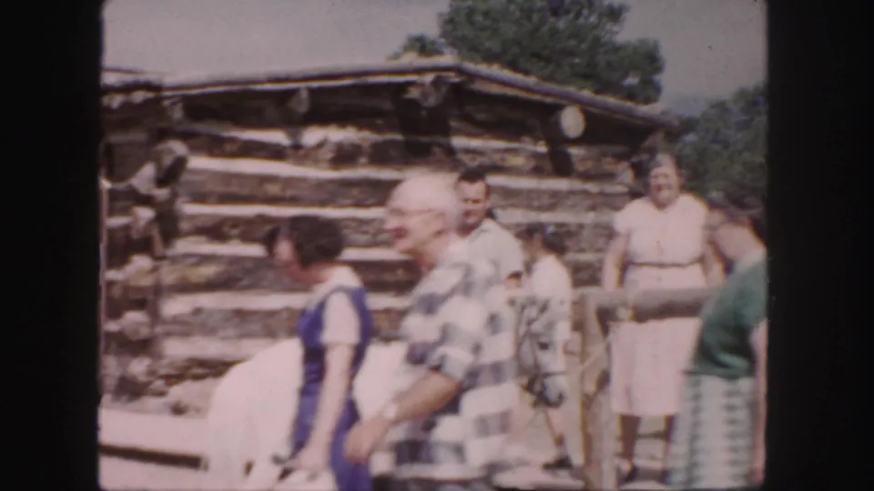1958 Colorado home-movie frame with several visitors near a white horse and a log cabin, with soft focus and faded color.