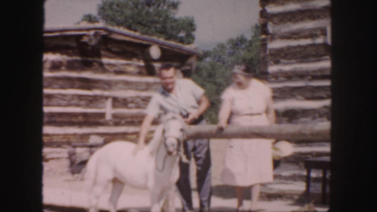 1958 Colorado home-movie frame showing a man with a small white pony and a woman near weathered log structures.