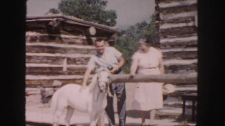 1958 Colorado home-movie frame showing a man with a small white pony and a woman near weathered log structures.