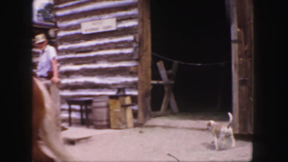 1958 Colorado home-movie frame with a small dog in front of an open log-building entrance and nearby ranch objects.