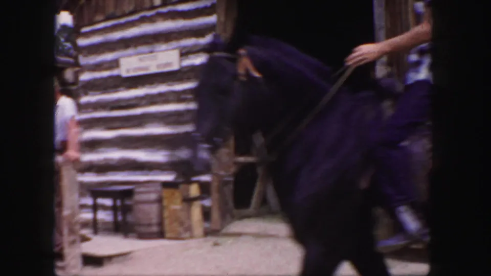 1958 Colorado home-movie frame showing a young-looking rider on a dark horse beside rustic log buildings.