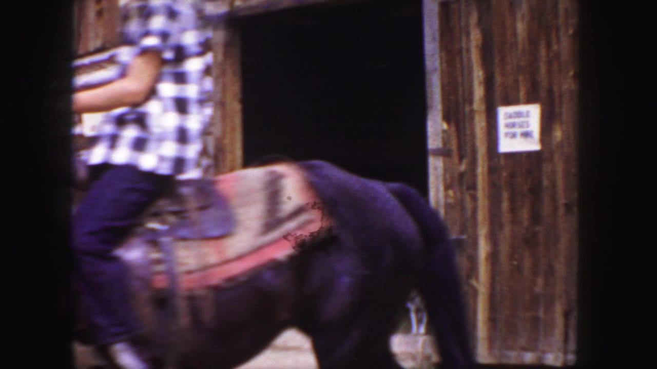 In Colorado home-movie footage from 1958, a boy in plaid rides a saddled dark horse past a log barn entrance.