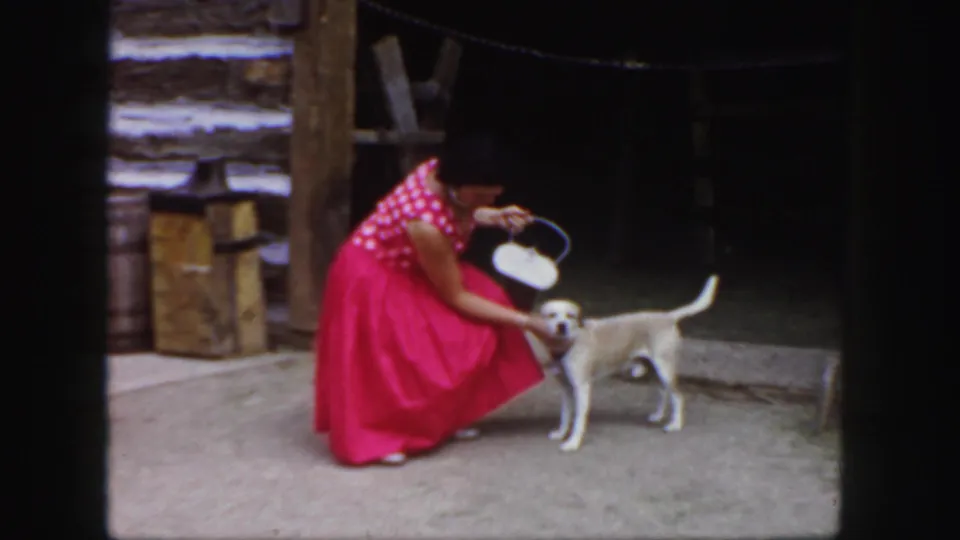 The 1958 Colorado home-movie frame shows a woman crouched beside a small dog at a dark barn entrance.