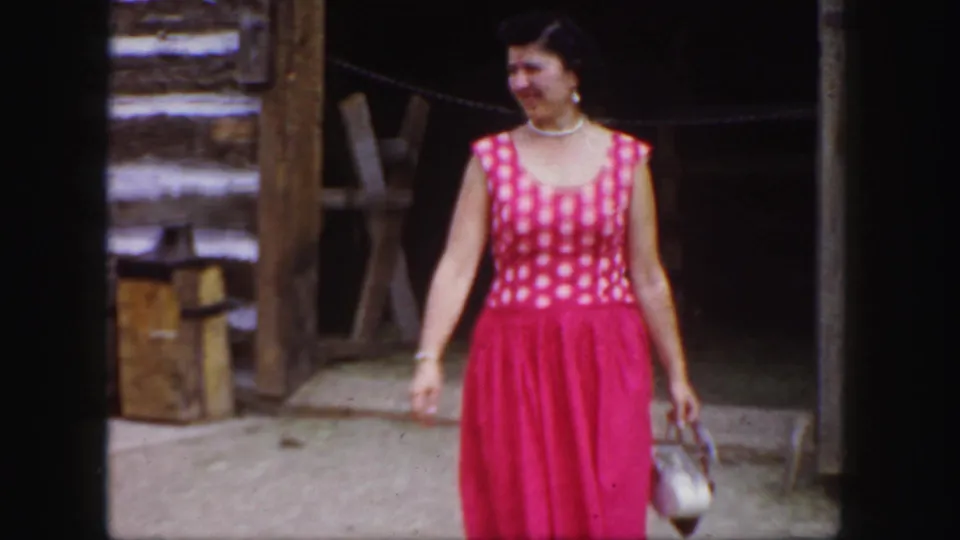 A woman in a bright red polka-dot dress smiles near the rustic Colorado cabin doorway, with her white handbag at her side.