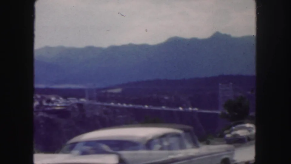 Home-movie view of a mountain valley and bridge with a mid-century car moving through the foreground.