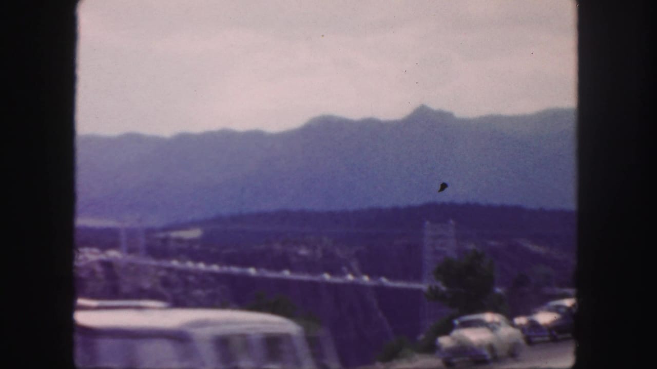 1958 Colorado home-movie frame showing classic cars by a canyon road, a long suspension bridge, and blue mountain ridges in the distance.