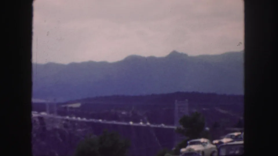 1958 Colorado frame with a canyon suspension bridge, roadside cars, dark foreground framing, and distant mountain peaks under a pale sky.