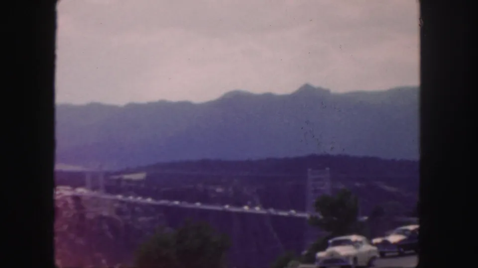 Faded 8mm Colorado view of a canyon suspension bridge, low hills, distant mountains, and roadside vegetation, framed by dark film edges.