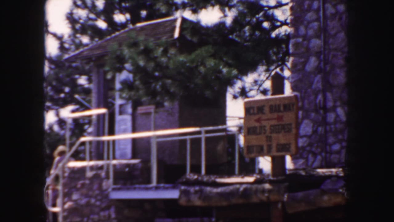 1958 Colorado home-movie frame showing a yellow Incline Railway sign near a stone building, white railing, trees, and a small visitor figure.