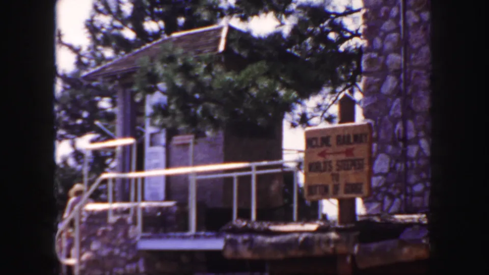 Colorado 1958 home-movie frame showing the Incline Railway sign, rustic stone building, white railing, trees, and a visitor near the path.