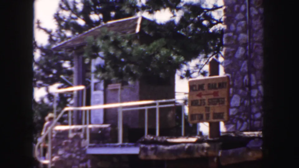 Final 1958 Colorado frame of a yellow Incline Railway sign at a mountain park, with stone architecture, white railing, trees, and home-movie grain.