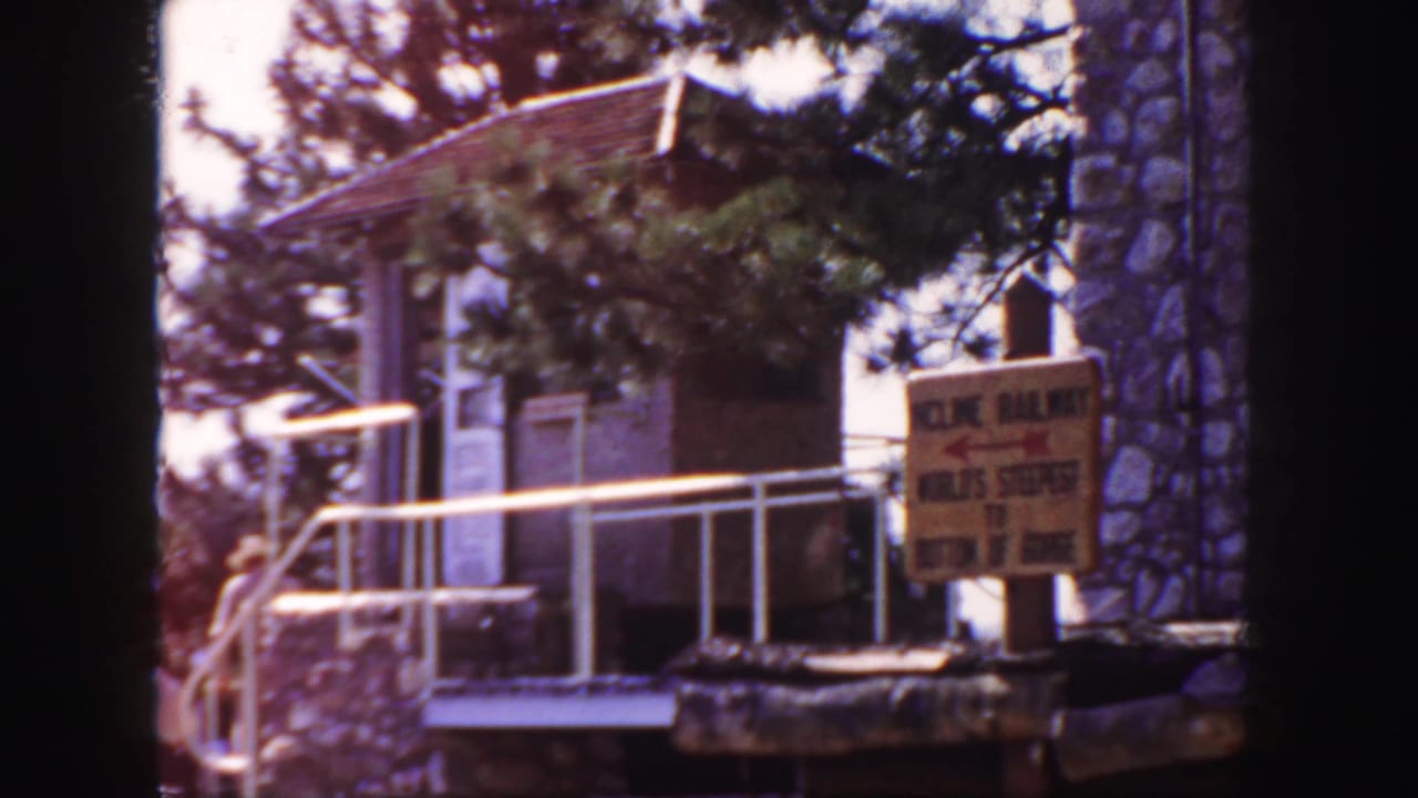 1958 Colorado home-movie frame showing a stone mountain station, ramp railings, trees, and an incline railway sign near the entrance.