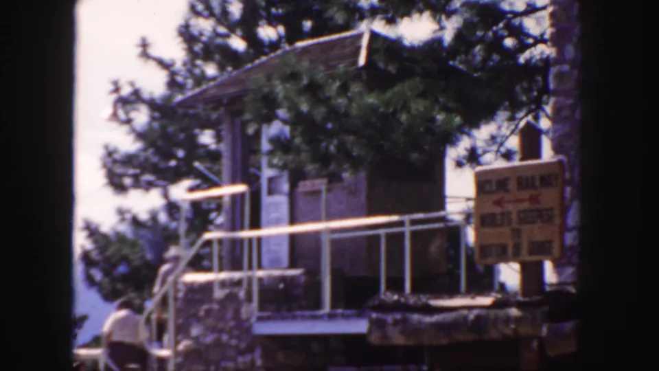 1958 Colorado frame with a rustic incline railway station, posted sign, white railings, trees, and a visitor partly visible near the ramp.