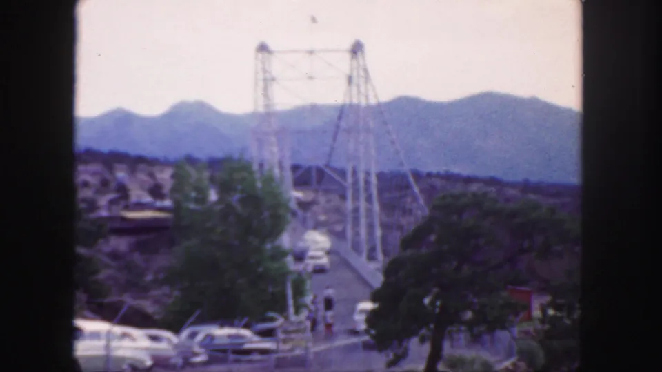 1958 Colorado home-movie view of a suspension bridge over canyon terrain, with vintage cars below and hazy mountains beyond.