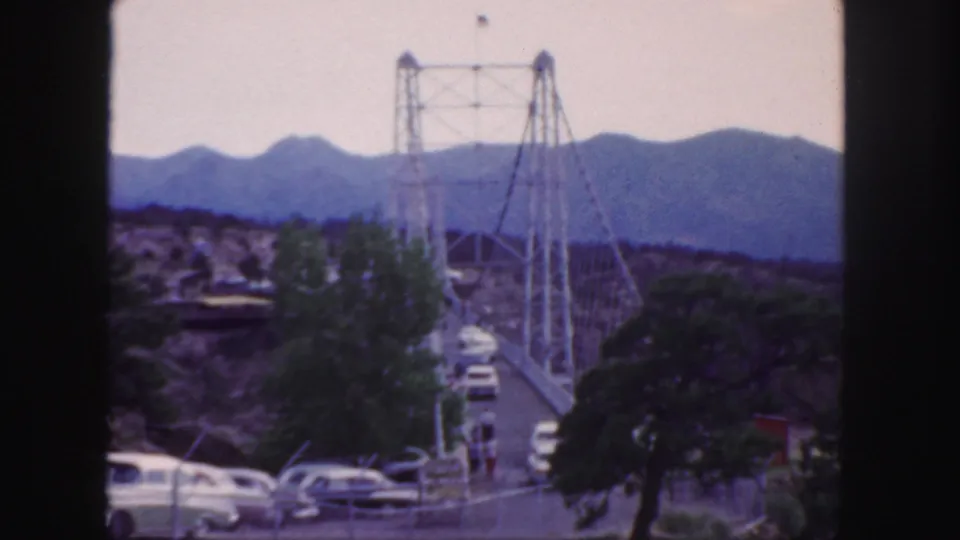 1958 Colorado frame showing a canyon suspension bridge, mid-century cars, sparse trees, rocky terrain, and mountain ridges in the background.