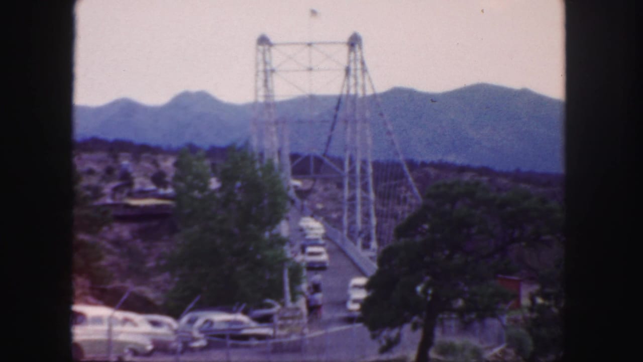 1958 Colorado 8mm frame showing mid-century cars approaching and crossing a suspension bridge backed by mountain ridges.
