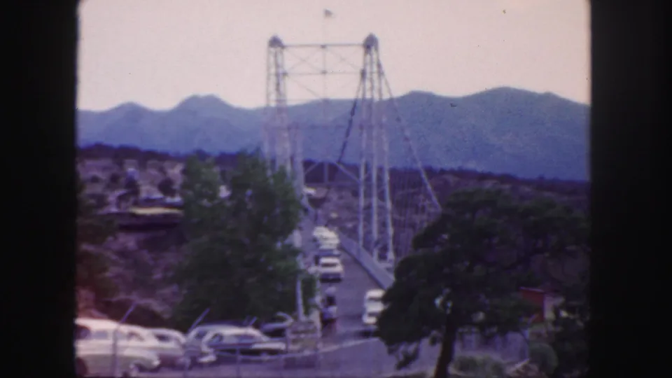 1958 Colorado 8mm frame showing mid-century cars approaching and crossing a suspension bridge backed by mountain ridges.