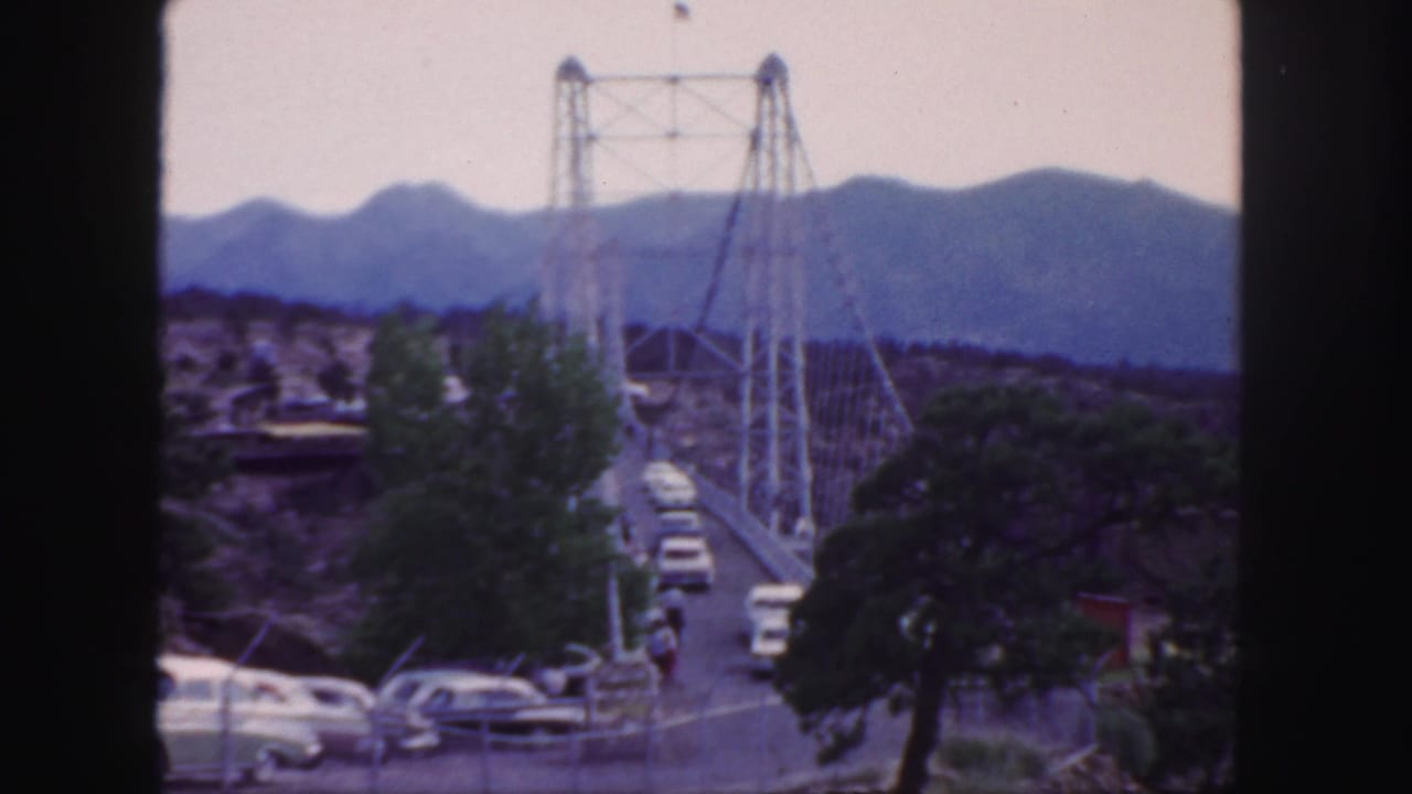 Cars cross a suspension bridge with trees and mountains beyond.