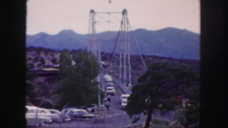 Mid-century cars travel across a Colorado suspension bridge in a soft 8mm frame with trees and distant mountains.
