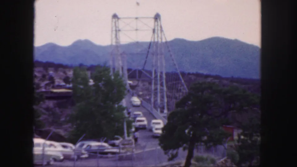 8mm Colorado frame with cars on and near a suspension bridge, roadside trees, and blue mountain ridges in the distance.