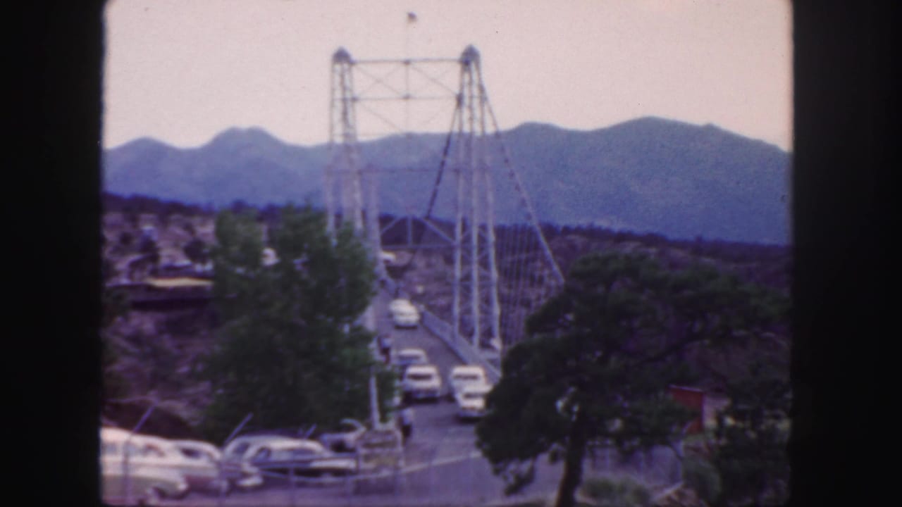 1958 Colorado 8mm view of a suspension bridge carrying cars across a canyon area with mountains in the distance.
