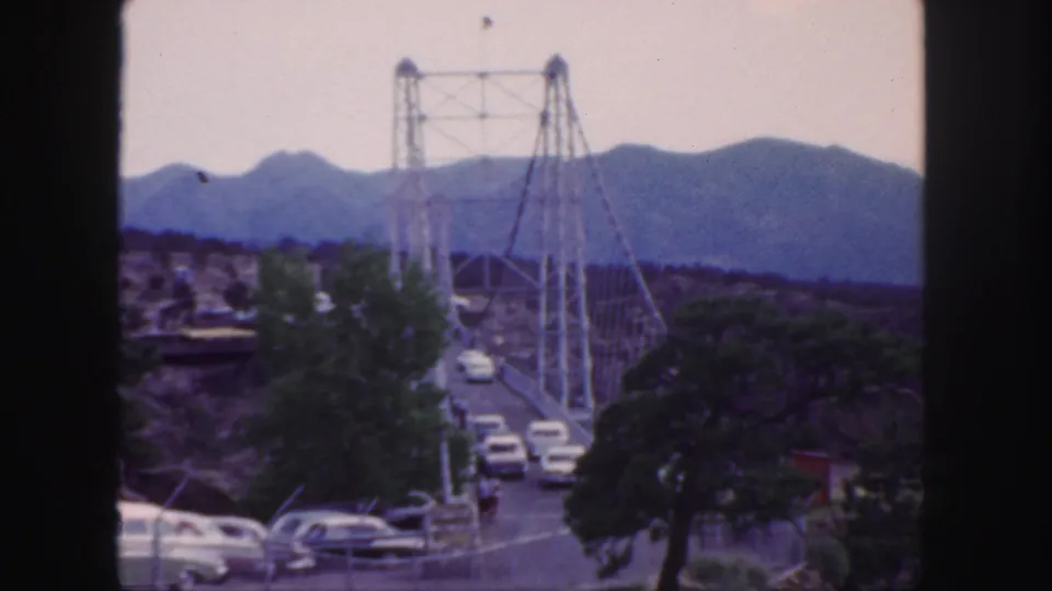 Distant 1958 Colorado travel view of cars on a suspension bridge, framed by trees and a hazy mountain backdrop.