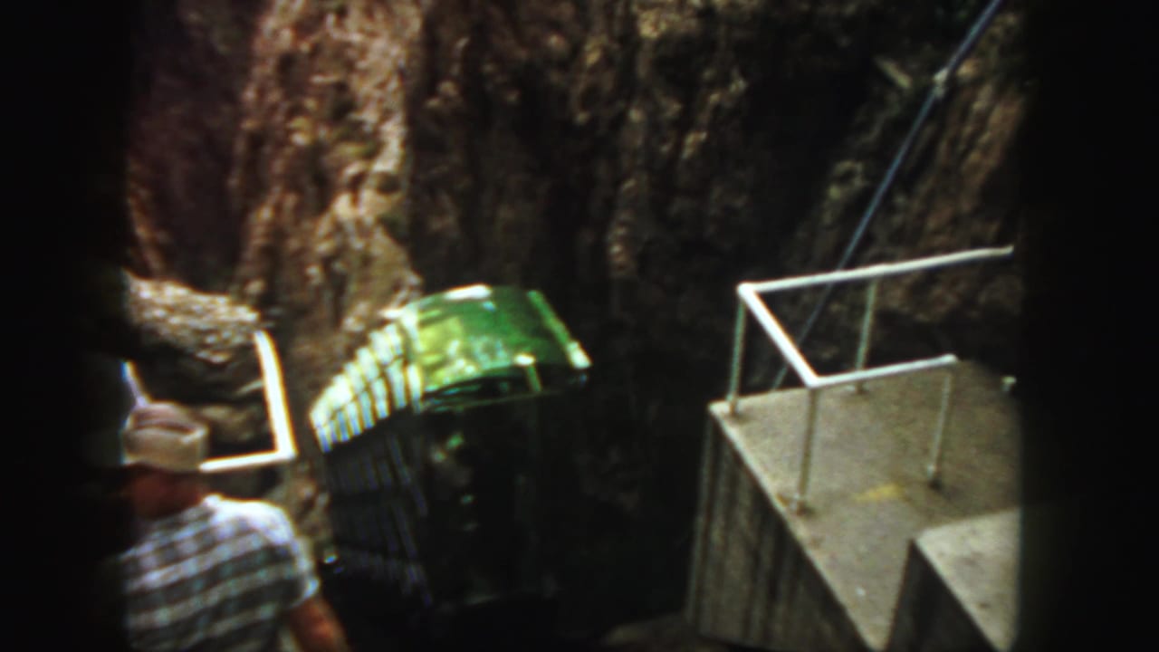 1958 Colorado 8mm frame showing a green rail bus ascending a steep canyon track near a concrete platform with an adult spectator.