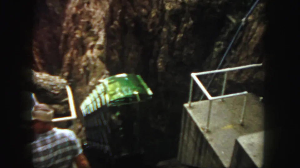 1958 Colorado 8mm frame showing a green rail bus ascending a steep canyon track near a concrete platform with an adult spectator.
