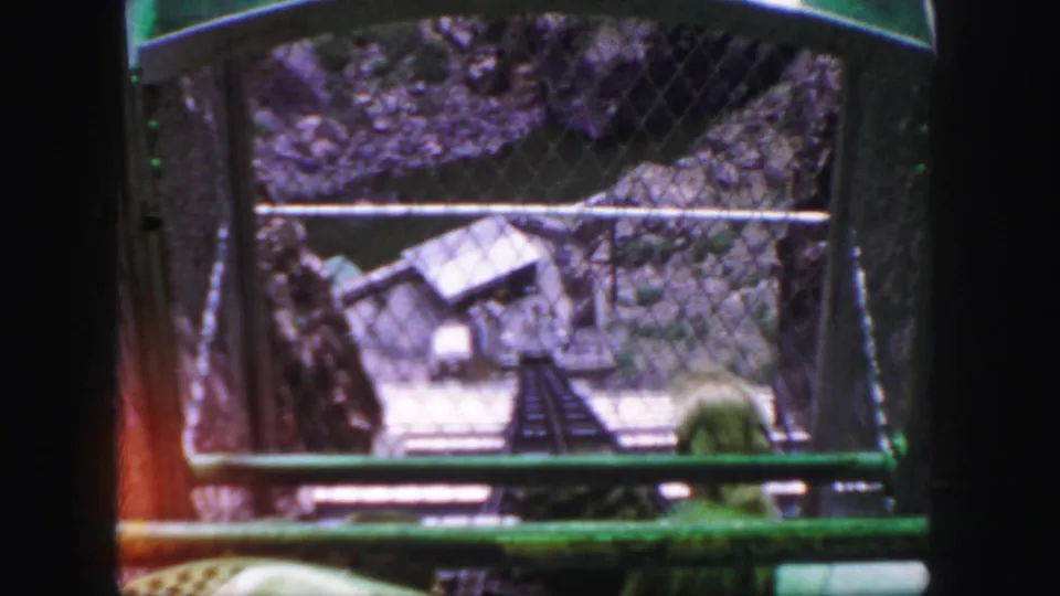 The mountain railway car looks past chain-link fencing toward a narrow track and a small structure below in the canyon.