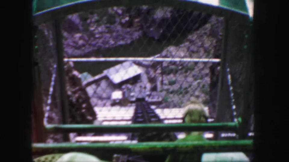 A later frame repeats the descending railway view through chain-link fencing, with rocky canyon walls and green car rails visible.
