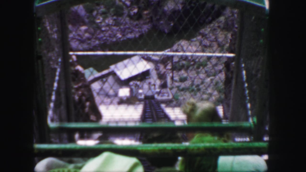 1958 Colorado 8mm frame from inside a green cable car, looking through chain-link fencing toward a steep track and small lower structure.