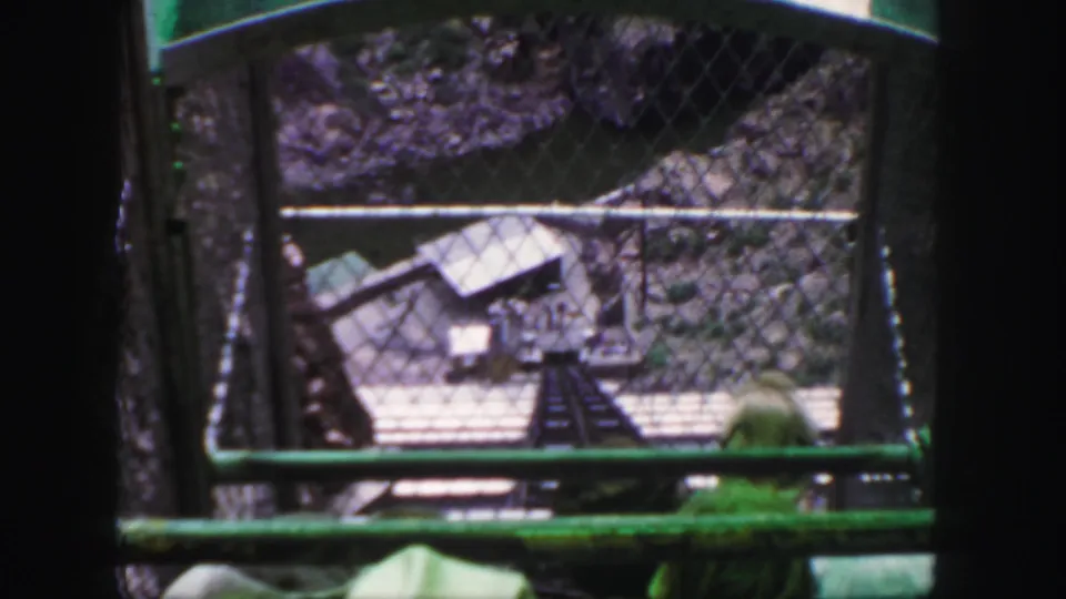 A home-movie frame looks from the moving cable car through fencing toward the narrow track and station-like structure below.